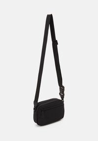 Sort slingtaske med en struktureret rem, lynlåslukket hovedrum og en frontlomme. Kompakt rektangulær form, der er velegnet til små nødvendigheder.