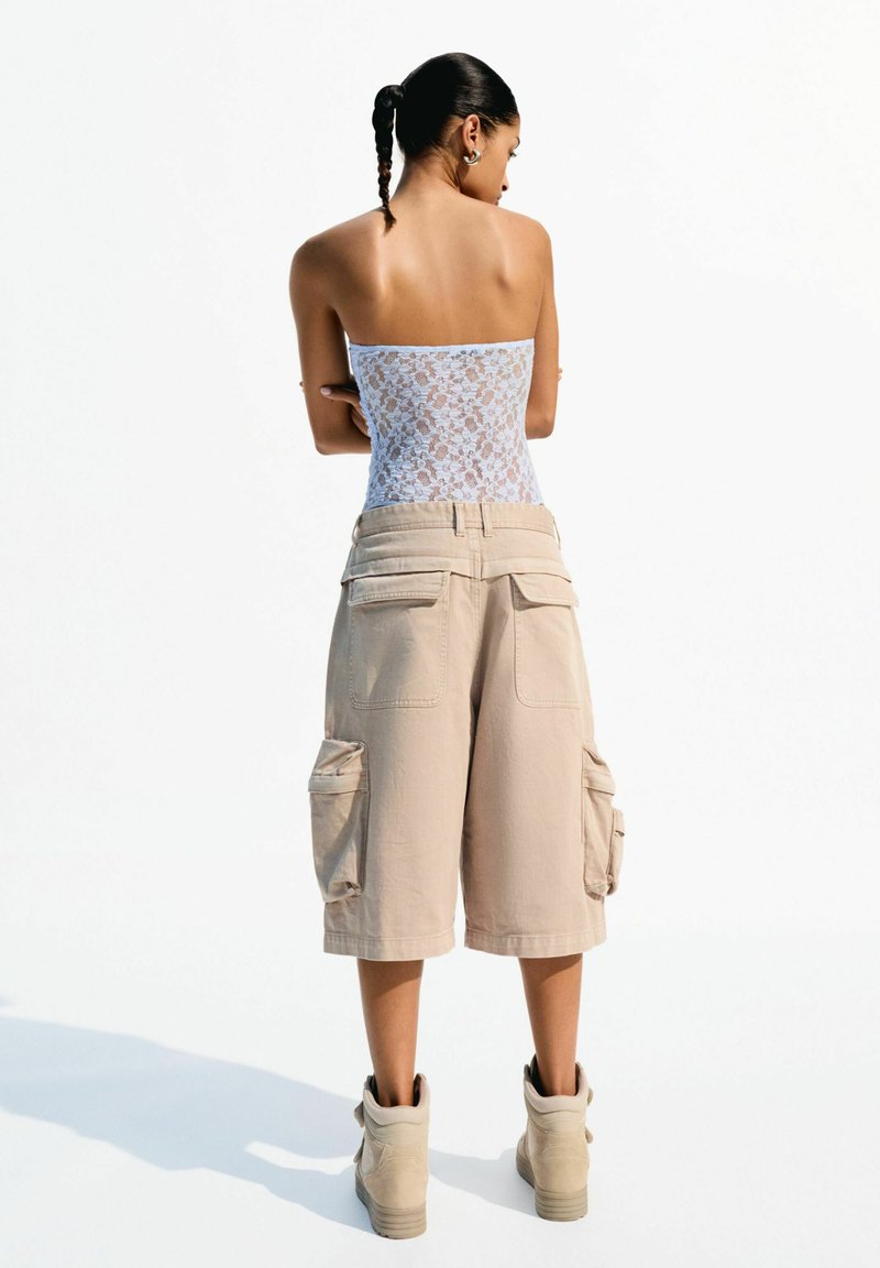 Bela čipkasta majica brez naramnic v kombinaciji z bež cargo hlačami z stranskimi žepi, oblečena z bež visokimi športnimi čevlji.