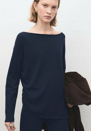 Haut bleu marine à manches longues avec un large col bateau et un tissu doux et texturé. Le mannequin tient une veste marron, sur un fond neutre.