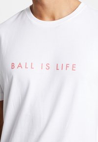 Camiseta de algodón blanca con cuello redondo y mangas cortas. El texto del frente dice "BALONCESTO ES VIDA" en letras mayúsculas de color rosa suave.