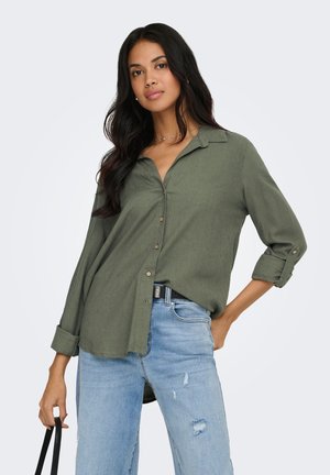 Mujer con el pelo largo y oscuro, vistiendo una camisa verde oliva abotonada y unos vaqueros azules claros rotos, sosteniendo la correa de un bolso negro.