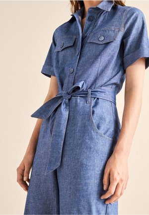 Denim jumpsuit in light blue with short sleeves, buttoned chest pockets, waist tie, and wide leg design, featuring a smooth texture.