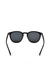 Lunettes de soleil à monture plastique noire avec des verres arrondis et des verres teintés foncés positionnées sur un fond blanc.