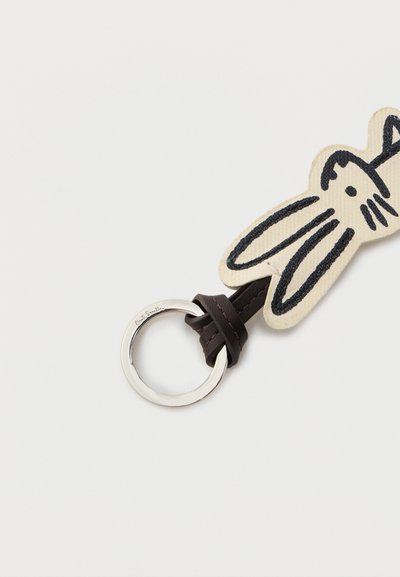 Porte-clés avec un anneau en argent attaché à une boucle en cuir foncé et une tête de lapin en tissu beige avec des traits du visage brodés en noir.