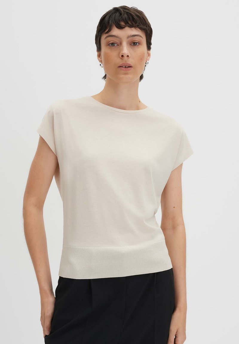 Top a maniche corte di colore crema con scollatura rotonda e orlo a costine. Tessuto morbido con una texture liscia. Indossato con pantaloni neri.