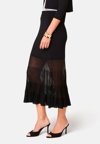 Schwarzer, plissierter Maxirock mit einem transparenten unteren Abschnitt, strukturiertem Stoff und einem ausgestellten Saum. Kombiniert mit schwarzen, offenen High Heels und einem goldenen Armband.