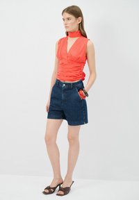 Top sin mangas rojo con un escote en V profundo y detalles fruncidos, combinado con pantalones cortos de mezclilla azules. Chanclas marrones con nudos como calzado.