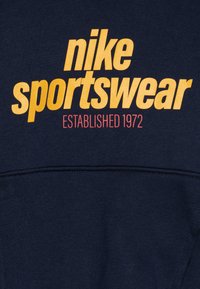 Jūras spēku sporta krekls ar lielu dzeltenu "nike sportswear" tekstu, mazāku sarkanu "ESTABLISHED 1972". Gluda auduma, ikdienas dizains, apaļa kakla izgriezums.