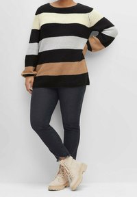 Persona che indossa un maglione a righe con bande nere, bianche e beige, jeans neri a vita alta con orli sfrangiati e stivali beige con lacci, in piedi.
