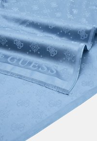 Svijetloplavi šal s glatkom teksturom, s diskretnim reljefnim uzorkom logotipa i resicama, koji prikazuje naziv branda "GUESS".
