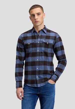 Uomo che indossa una camicia a quadri blu e nera con bottoni e due tasche sul petto, abbinata a jeans blu, in piedi contro uno sfondo chiaro e uniforme.