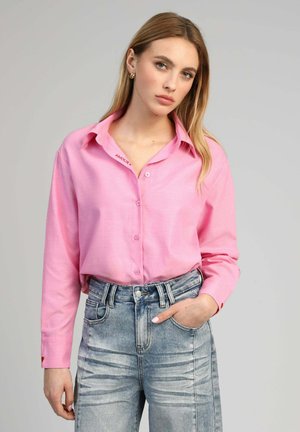 Jeune femme aux longs cheveux blonds portant une chemise rose boutonnée rentrée dans un jean bleu délavé taille haute, debout devant un fond uni.