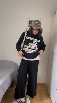 Άτομο που φοράει καπέλο καμουφλάζ, μαύρο φούτερ με λευκό ισπανικό κείμενο, φαρδιά μαύρη παντελόνα, λευκά αθλητικά παπούτσια και τσάντα με ιμάντα σχοινιού σε ένα υπνοδωμάτιο.