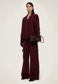 Bordowy garnitur z szerokimi spodniami i dopasowaną marynarką. Modelka trzyma małą czarną torebkę, nosi czarne rękawiczki. Gładka tkanina, minimalistyczne detale.