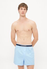 Șorturi de baie de culoare albastru deschis, fabricate dintr-un material neted, având o talie cu logo-ul Lacoste în culori alb-negru și un sistem de închidere cu un singur buton.