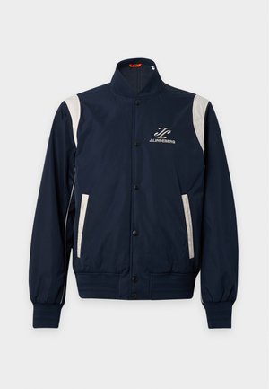 Jeune homme portant une veste bomber bleu marine avec des accents blancs, un pull foncé, un jean bleu clair, une ceinture noire et des baskets blanches, debout sur un fond uni.