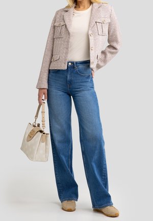 Vrouw draagt een beige gestructureerd jasje, wit topje, blauwe high-waisted jeans, beige sneakers, houdt een beige handtas met patroon naast zich.