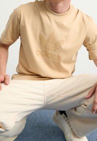 Camiseta beige de manga corta con logo en relieve de Adidas y diseño de hojas. Material suave de algodón. Combinada con pantalones beige y zapatillas blancas.