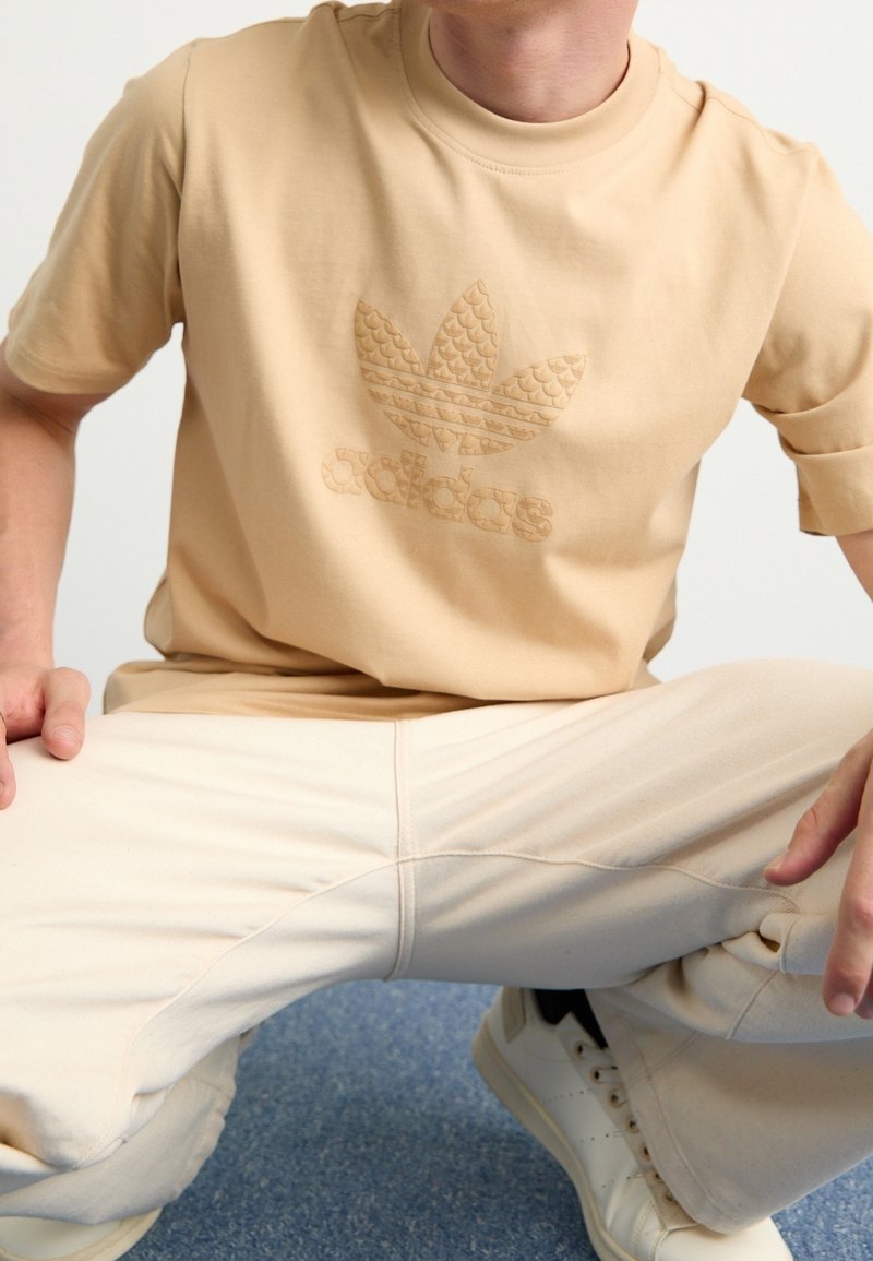 Camiseta beige de manga corta con logo en relieve de Adidas y diseño de hojas. Material suave de algodón. Combinada con pantalones beige y zapatillas blancas.