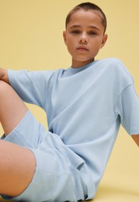 Niño con el pelo corto que lleva puestos pantalones cortos texturizados azul claro y una camiseta, sentado frente a un fondo liso de color amarillo pálido.