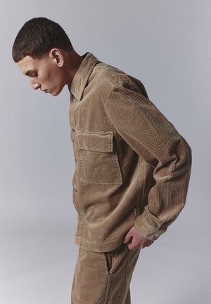 Brown corduroy shirt jacket with a collar, two chest pockets, and button closure. Features a textured surface and relaxed fit design.