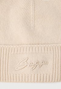 Căciulă tricotată în bej deschis cu manșetă cu dungi, având un logo brodat subtil. Textură moale și design simplu.