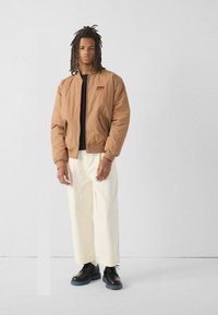 Bruine bomberjack met een textuurafwerking, voorzien van een ritssluiting aan de voorkant, zijzakken en een rood logo detail. Gecombineerd met roomkleurige broek met wijde pijpen en zwarte schoenen.