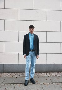Sort blazer over en lys blå knap-op skjorte, parret med lys blå jeans og sorte sko, stående mod en dæmpet rektangulær væg.
