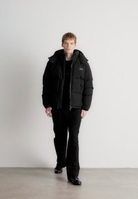 Crna zimska jakna s patentnim zatvaračem sprijeda, kapuljačom i detaljem sa zakrpom. Nosi se preko crne majice i u kombinaciji je s crnim hlačama i cipelama.