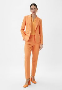 Maßgeschneiderter orangefarbener Hosenanzug mit einem Ein-Knopf-Blazer und schmal geschnittenen Hosen. Paar orangefarbener Blockabsatzschuhe. Glatte, einheitliche Textur.