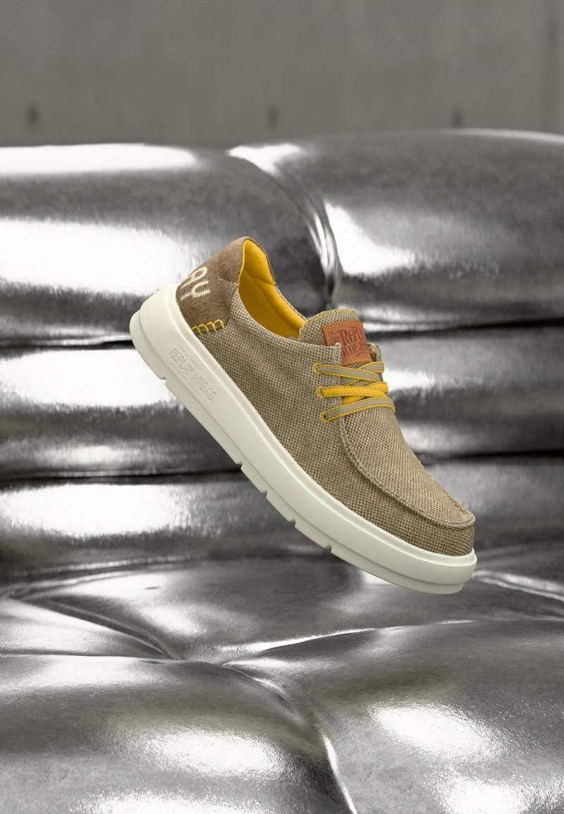 Μπεζ καμβαδένιο παπούτσι με κίτρινα κορδόνια και λεπτομέρειες, διαθέτοντας υφή στην επάνω επιφάνεια, λευκή σόλα και καφέ λογότυπο.