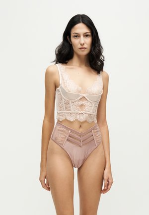 Femme aux cheveux foncés portant un haut de lingerie en dentelle rose clair et un bas taille haute assorti, debout devant un fond uni.