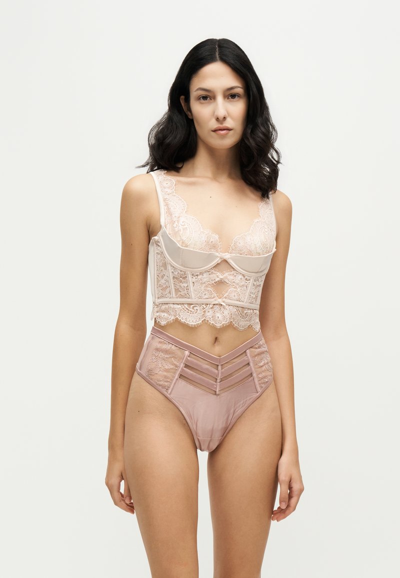 Femme aux cheveux foncés portant un haut de lingerie en dentelle rose clair et un bas taille haute assorti, debout devant un fond uni.