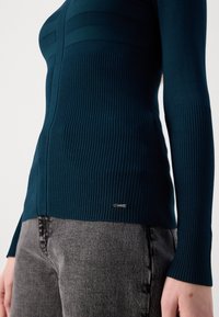Gros plan d'une personne portant un pull en maille côtelée vert sarcelle foncé et un jean noir délavé, montrant la texture et un petit logo métallique sur l'ourlet du pull.