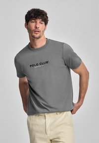 Camiseta de algodón gris con cuello redondo, que presenta el logo "POLO CLUB" y el año de fundación impresos en negro. Textura suave, corte relajado.