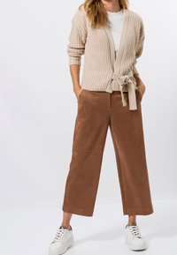 Beige pletený kardigan s páskem a žebrovaným lemem, nošený přes bílý top. Hnědé kalhoty s širokými nohavicemi z měkké tkaniny, doplněné bílými teniskami.