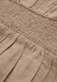 Lys beige stof med et tekstureret smock-mønster i midten, der har samlede sektioner langs kanterne, hvilket skaber en blød fald.