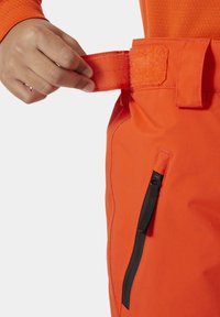 Světle oranžové kalhoty s nastavovacím pasem na suchý zip a černou zipovou kapsou na boku. Hladká textilie a přiléhavý střih.