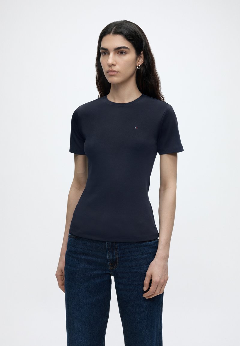 Νεαρή γυναίκα με μακριά σκούρα μαλλιά, ντυμένη με στενό μπλε σκούρο κοντομάνικο μπλουζάκι και σκούρο μπλε τζιν, στέκεται μπροστά σε ένα απλό ανοιχτό φόντο.