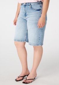 Bermudas en denim bleu clair avec une coupe droite, dotées d'une taille mi-haute, de cinq poches et d'un ourlet effiloché. Portées avec des tongs.