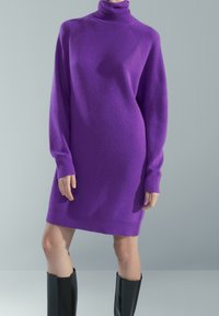 Robe col roulé en maille violette avec manches longues, offrant une coupe décontractée et des poignets côtelés, portée avec des bottes noires montantes jusqu'au genou.