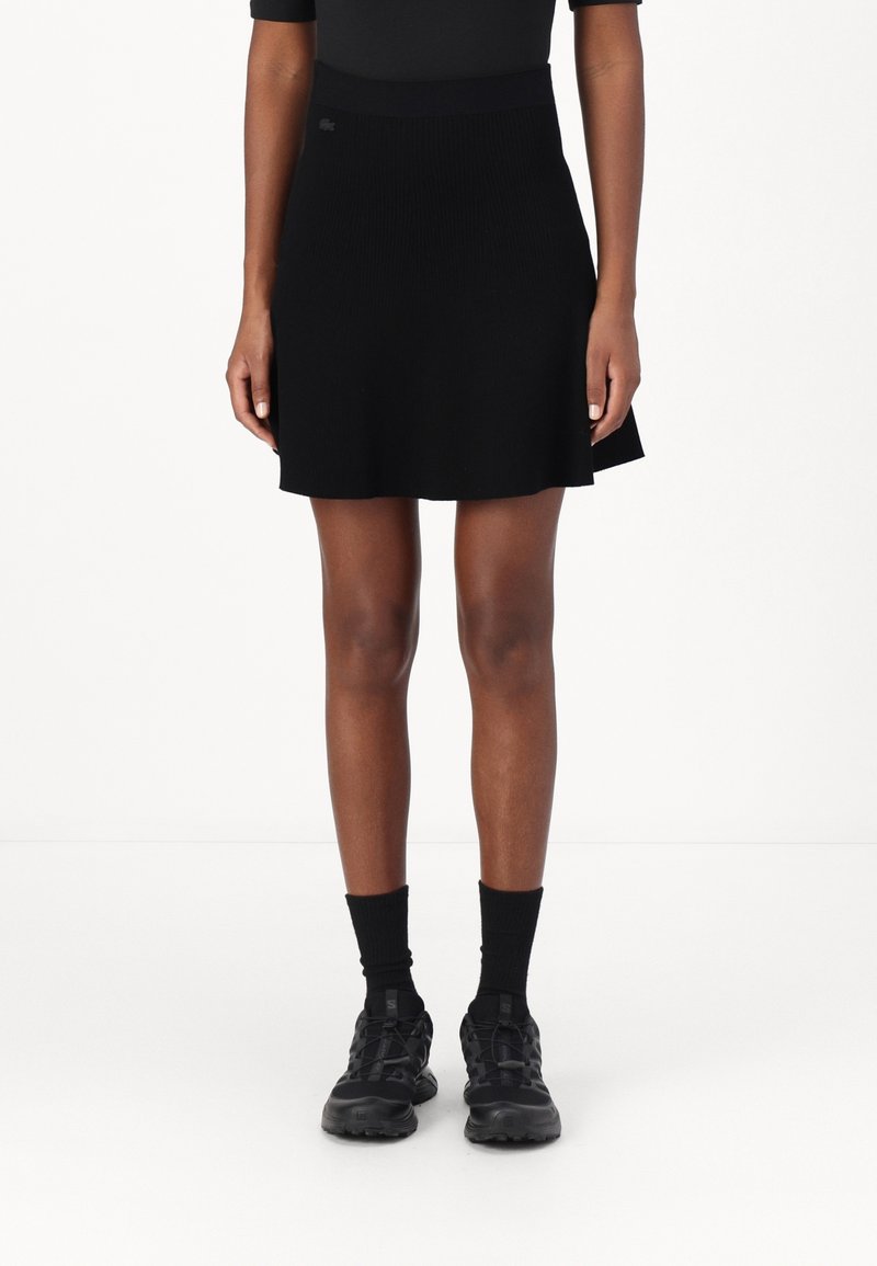 Falda negra de punto acanalado con cintura ceñida, con dobladillo ligeramente acampanado, combinada con zapatillas deportivas negras y calcetines largos negros.