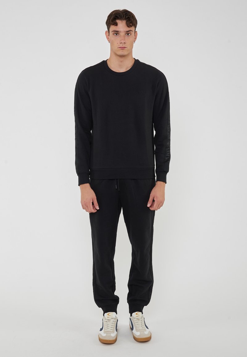 Felpa nera con maniche lunghe e polsini a coste, abbinata a pantaloni della tuta neri e sneakers bianche con dettagli blu navy. Fit semplice e rilassato.