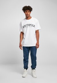 Camiseta blanca de manga corta con "SOUTHPole ORIGINALS" impreso en negro, combinada con pantalones de mezclilla azules y zapatillas deportivas blancas de caña alta.