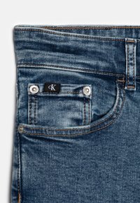 Jeans in denim con una lavatura blu scuro, caratterizzati da una piccola tasca frontale, accessori in argento e un'etichetta nera con scritte bianche.