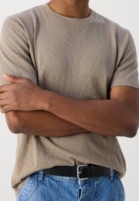 T-shirt corto a maniche corte color marrone chiaro con collo a giro, texture morbida, con branding discreto. Abbinato a jeans blu e una cintura nera.