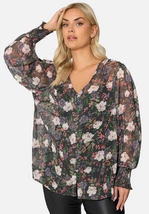 Donna con capelli biondi che indossa una blusa nera trasparente con motivo floreale nei toni del rosa e viola e polsini arricciati, mentre posa con una mano sulla testa.