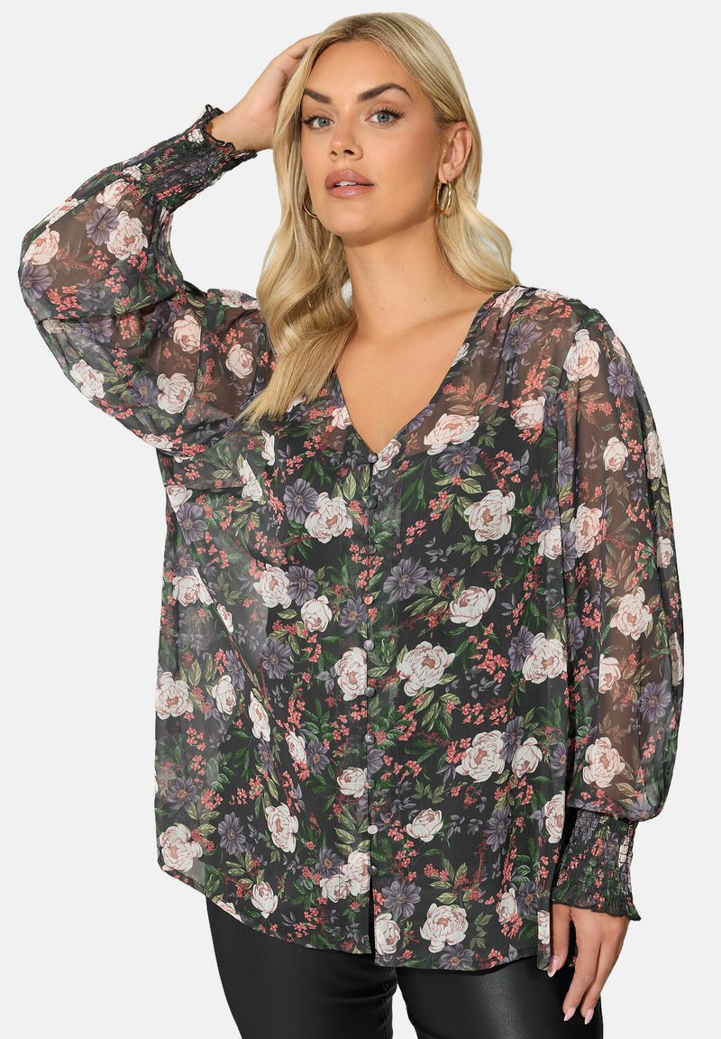Donna con capelli biondi che indossa una blusa nera trasparente con motivo floreale nei toni del rosa e viola e polsini arricciati, mentre posa con una mano sulla testa.