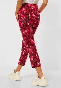 Person klädd i röd-och-svart ormskinnsmönstrade cropped byxor med fickor och vita chunky sneakers, stående mot en enkel bakgrund.