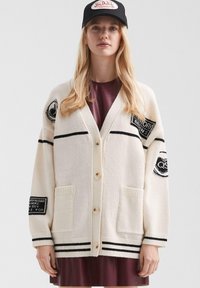 Młoda kobieta ubrana w kremowy kardigan w czarne paski i łaty, nosząca burgundową sukienkę oraz czapkę Von Dutch w kolorze czarnym.